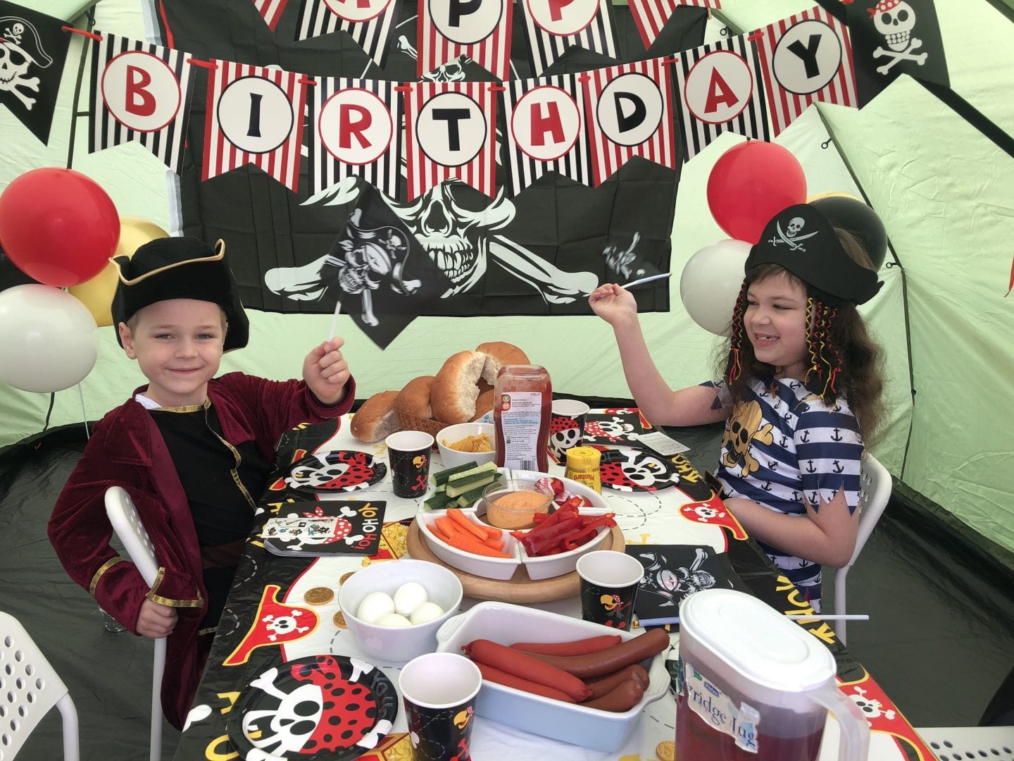 2 children sat at a table of food, waving small pirate flags, in a tent decorated for a pirate party