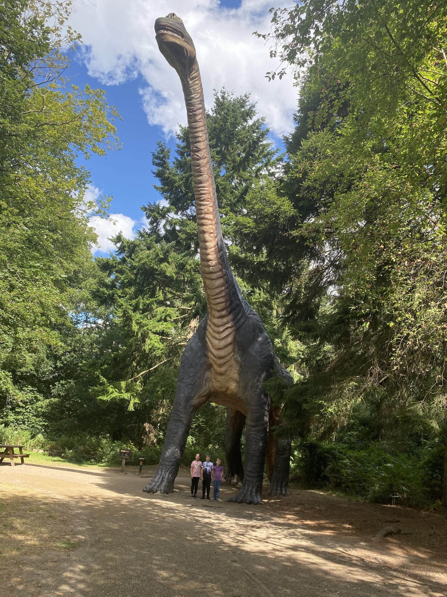 3 children standing under a large statue of a diplodocus