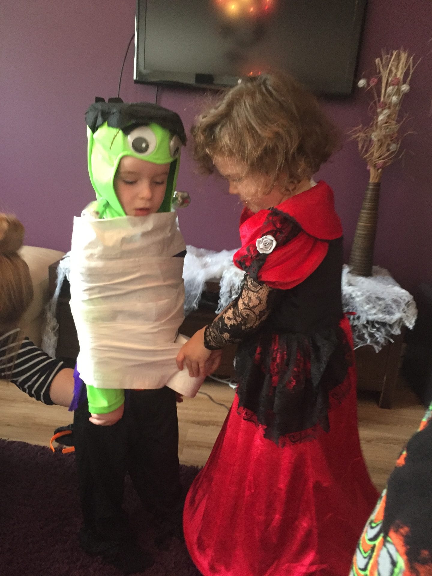 2 small children dressed in Halloween costums, one is wrapping the other in toilet paper